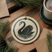 a ceramic ornament with a black and white swan design, placed on a wooden surface alongside a cup of coffee and some pine branches.
