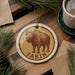 A circular ornament featuring a bison illustration and the word "Takin" on a wooden surface, surrounded by pine branches and a cup of coffee.