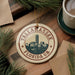 A circular ornament with the text "TALLAHASSE FLORIDAUA" and a graphic of a city skyline, hanging on a branch with pine needles.
