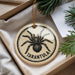 A circular ornament with a black and white illustration of a tarantula, surrounded by a gold string.