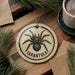 A decorative ornament featuring a black and white illustration of a tarantula, with the word "Tarantula" written around the center of the image. The ornament is hanging from a string, and there is a cup of coffee and some pine branches in the background.
