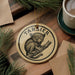 A circular ornament featuring a monkey with large eyes, hanging on a wooden surface next to a cup of coffee and some pine branches.