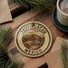 A circular ornament with the text "TEOTIHUACAN MEXICO" printed on it, surrounded by pine branches and a cup of coffee on a wooden surface.