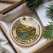 A round ornament with a design of Theodore Roosevelt National Park, featuring a mountain landscape and a river, is displayed in a box with pine branches.