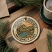 A circular ornament with the text "THEODORE ROOSEVELT NATIONAL PARK USA" is displayed on a wooden surface, surrounded by pine branches and a cup of coffee.