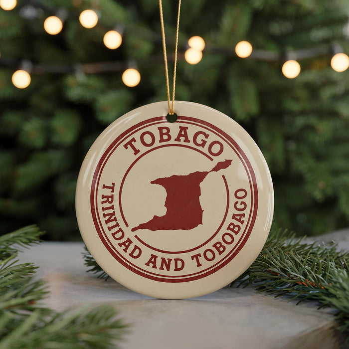 A circular ornament with the text "TOBAGO TRINIDAD AND TOBAGO" printed on it, along with a red outline of the island of Trinidad and Tobago. The ornament is hanging from a string, and there are pine branches visible in the background.