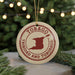 A circular ornament with the text "TOBAGO TRINIDAD AND TOBAGO" printed on it, along with a red outline of the island of Trinidad and Tobago. The ornament is hanging from a string, and there are pine branches visible in the background.