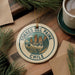 A decorative ornament with the text "TORRES DEL PAINE CHILE" on it, featuring a mountain landscape design, is placed on a wooden surface alongside a cup of coffee and some pine branches.