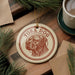 A round ornament with a dog's head and the text "TOSA DOG" is displayed on a wooden surface, surrounded by pine branches and a cup of coffee.