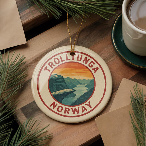 A circular ornament with the text "TROLLUNGA NORWAY" and an illustration of a mountain landscape hangs on a wooden surface, surrounded by pine branches and a cup of coffee.