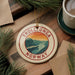 A circular ornament with the text "TROLLUNGA NORWAY" and an illustration of a mountain landscape hangs on a wooden surface, surrounded by pine branches and a cup of coffee.