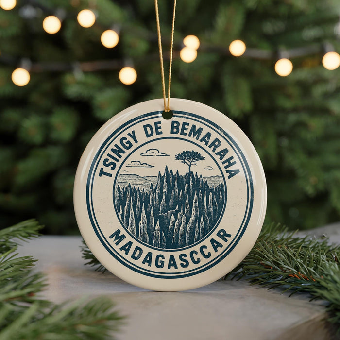 A circular ornament with a graphic design featuring a mountain landscape and the text "Tsingy de Bemaraha Madagascar" is displayed on a table with pine branches in the background.
