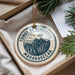 A circular ornament with a tree design and text, placed in a box with pine branches.