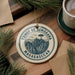 A circular ornament with the text "TSINGY DE BEMARAH" and "MADAGASCAR" printed on it, surrounded by pine branches and a cup of coffee on a wooden surface.