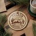 A circular ornament with the text "TUCSON ARIZONA US" printed on it, surrounded by pine branches and a cup of coffee on a wooden surface.