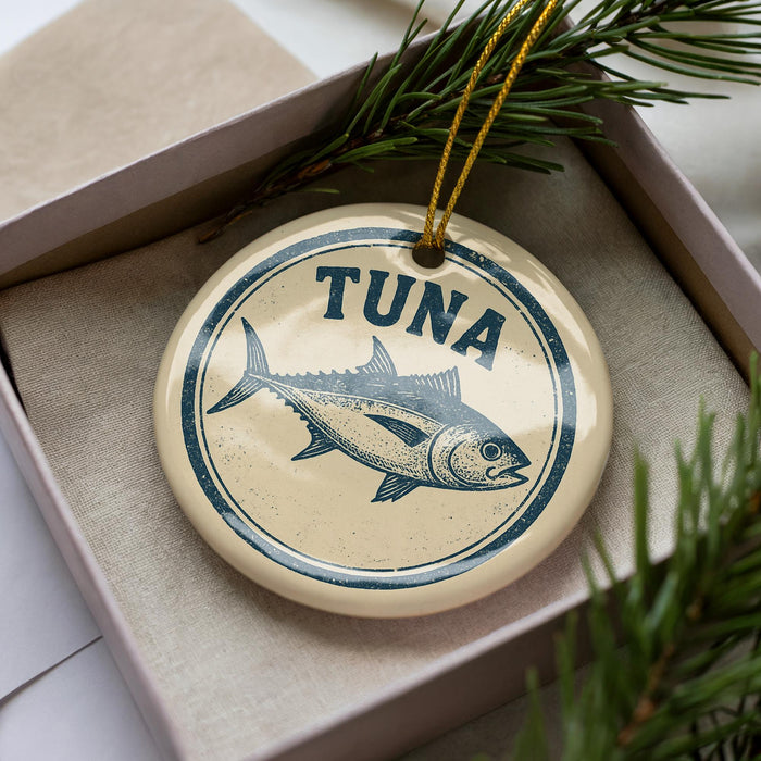A round ornament with a blue and white design featuring a fish, specifically a tuna, is displayed in a box with pine branches.