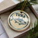A round ornament with a blue and white design featuring a fish, specifically a tuna, is displayed in a box with pine branches.