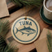 A round ornament with the word "Tuna" printed on it, featuring a fish illustration, is placed on a wooden surface next to a cup of coffee and pine branches.