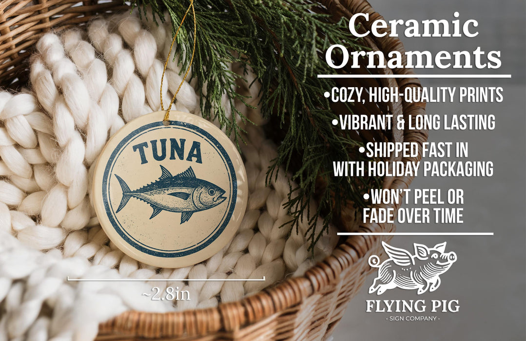 Ceramic ornaments, featuring a round ornament with a fish design and text, are displayed in a basket with pine branches.