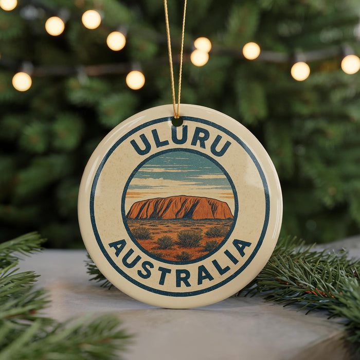 A circular ornament with the text "ULURU AUSTRALIA" and an illustration of Uluru, a famous rock formation in Australia, is displayed in front of a blurred Christmas tree with lights.