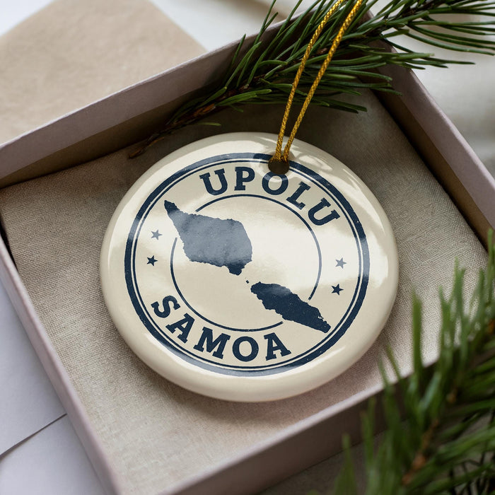 A circular ornament with the text "UPOLU SAMOA" printed on it, surrounded by a decorative border. The ornament is placed inside a box, with pine branches visible in the background.
