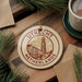 A circular ornament with the text "UTREcht Netherlands" on it, surrounded by pine branches and a cup of coffee on a wooden surface.