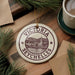 A decorative ornament with the text "VICTORIA SEYCHELLES" and an illustration of a town or village, placed on a wooden surface alongside a cup of coffee and some pine branches.