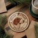 A circular ornament featuring a Welsh Springer Spaniel dog is displayed on a wooden surface, surrounded by pine branches and a cup of coffee.