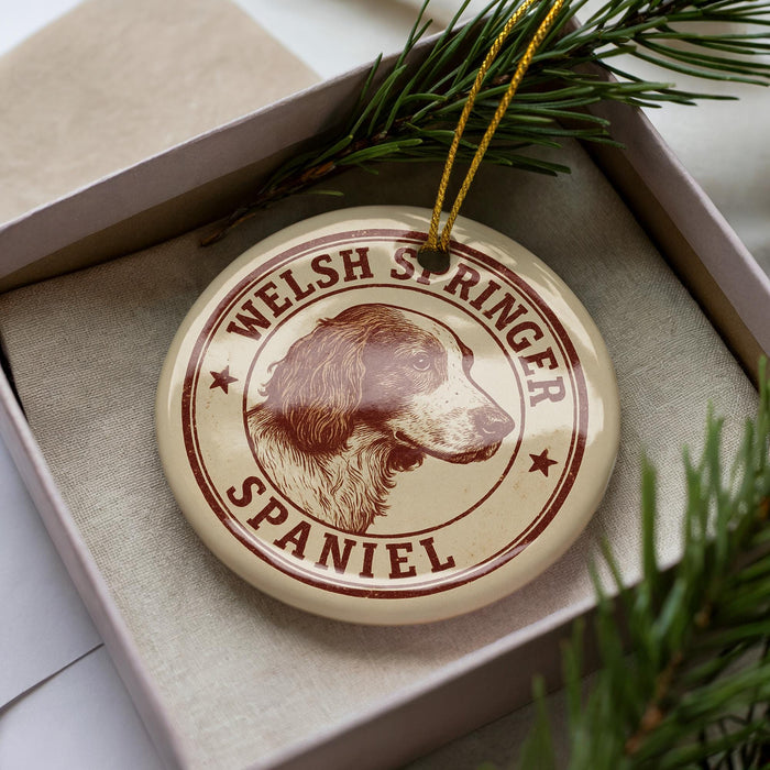 A circular ornament featuring a Welsh Springer Spaniel dog's head, with the text "WELSH SPRINGER SPANIEL" printed on it. The ornament is placed inside a box, surrounded by pine branches.