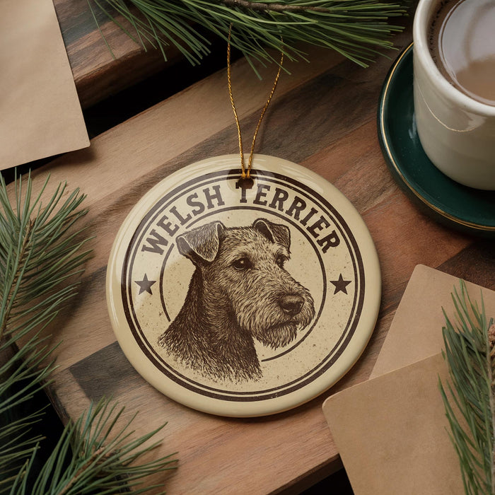 A circular ornament featuring a Welsh terrier dog's head, with the text "WELSH TERRIER" printed on it, is displayed on a wooden surface alongside a cup of coffee and some pine branches.
