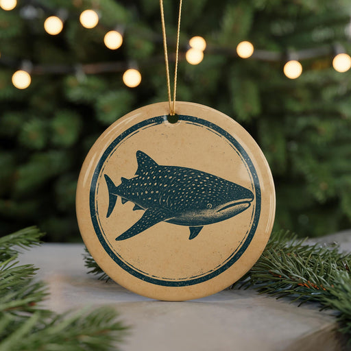 A round ornament with a blue and white design depicting a shark is hanging from a string, surrounded by pine branches and twinkling lights.