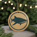 A round ornament with a blue and white design depicting a shark is hanging from a string, surrounded by pine branches and twinkling lights.
