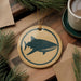 a round ornament with a blue and white design depicting a fish, placed on a wooden surface next to a cup of coffee and some pine branches.