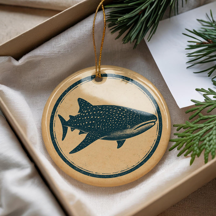 A round ornament with a blue and white design depicting a shark is placed in a box with pine branches.