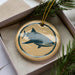A round ornament with a blue and white design depicting a shark is displayed in a box, surrounded by pine branches.