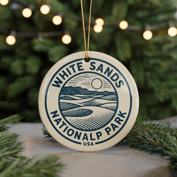A white ornament with the text "WHITE SAND'S NATIONALALP PARK USA" is displayed on a pine branch, with a blurred background of lights.