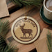 a decorative ornament with a deer illustration, placed on a wooden surface alongside a cup of coffee and some pine branches.