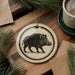 a ceramic ornament with a black and white illustration of a wild boar, hanging on a wooden surface.