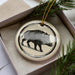 A ceramic ornament in the shape of a boar, with a black and white illustration of the animal, is displayed in a box with pine branches.