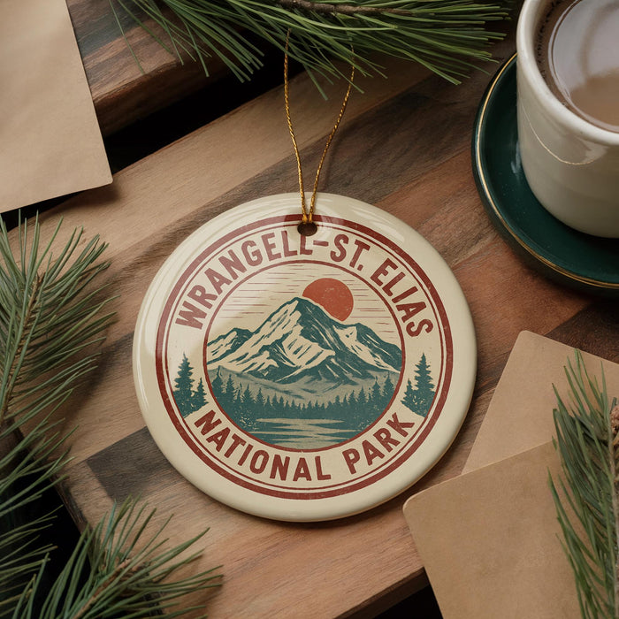 A circular ornament with the text "WRANGELAND-ST. ELIAS NATIONAL PARK" and a mountain landscape design hangs on a wooden surface, surrounded by pine branches and a cup of coffee.
