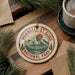 A circular ornament with the text "WRANGELAND-ST. ELIAS NATIONAL PARK" and a mountain landscape design hangs on a wooden surface, surrounded by pine branches and a cup of coffee.