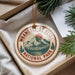 A circular ornament with a mountain and pine tree design, hanging from a string, placed on a white surface next to pine branches.