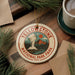 A decorative ornament featuring the Yellowstone National Park logo, surrounded by pine branches and a cup of coffee on a wooden surface.