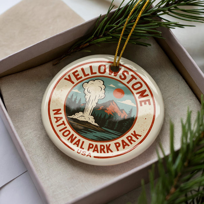 A decorative ornament in a box, featuring a circular design with a mountain landscape and a steam explosion, representing Yellowstone National Park.