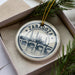 A circular ornament with a drawing of a bridge and the word "ZARAGOZA" on it, placed in a box with pine branches.
