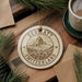 A decorative ornament with the text "ZERMATT SWITZERLAND" and a mountain illustration, placed on a wooden surface next to a cup of coffee and pine branches.