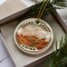 A decorative ornament in a box, featuring a mountain landscape with the text "Zhangyong Danxia China" printed on it.