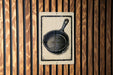 a black and white illustration of a frying pan on a wooden wall.