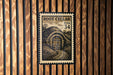 A vintage-style postage stamp featuring a wooden door and a landscape scene, mounted on a wooden wall.