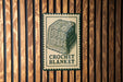 A sign with a crocheted blanket on it is displayed on a wooden wall.